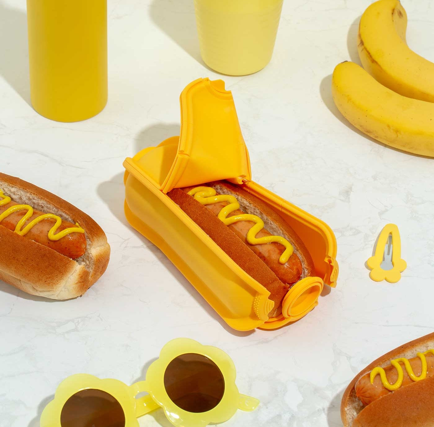 Sunshine Wrap'd Open Showing Hotdog With Mustard Inside Surrounded By Yellow Sunglasses and Hairclips and Bananas