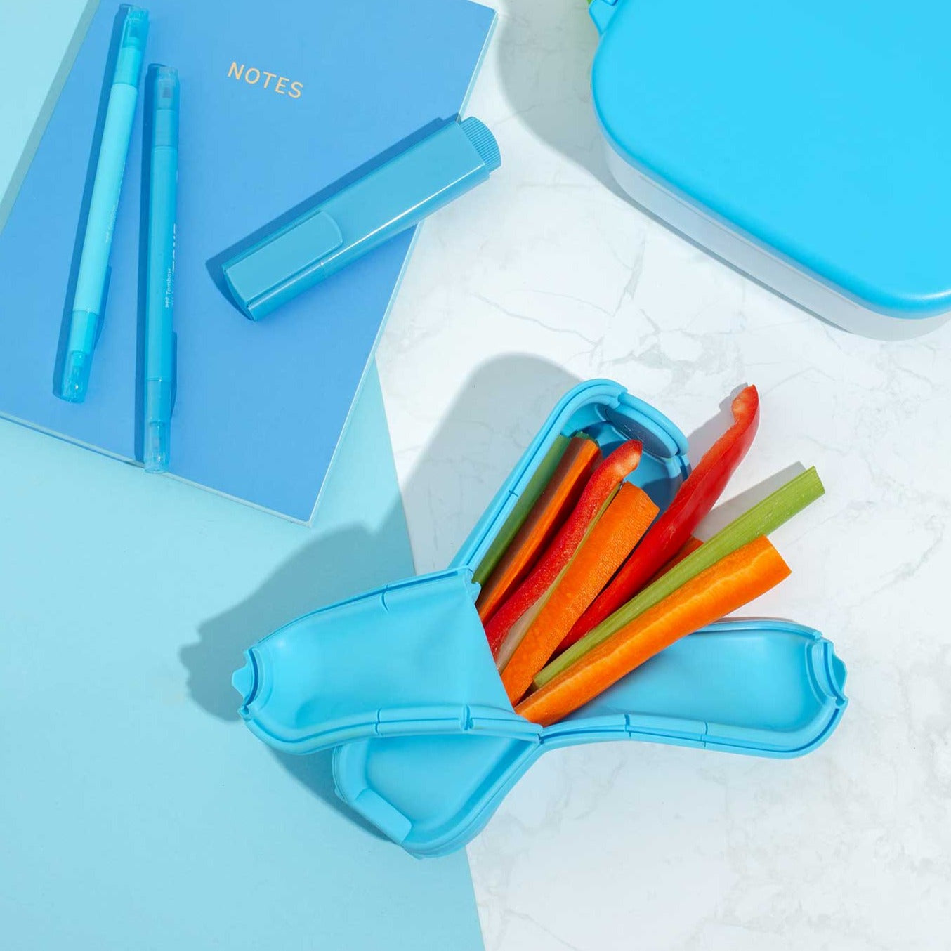 Blue Wrap'd Open Showing Vege Sticks Inside With Matching Blue Pens And Notebook Next To It