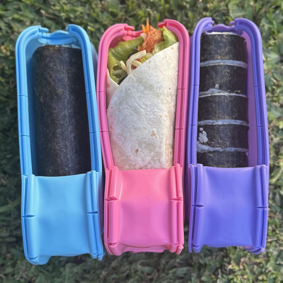 Three colourful Wrap'd wrap storage containers holding food items on a grassy background.