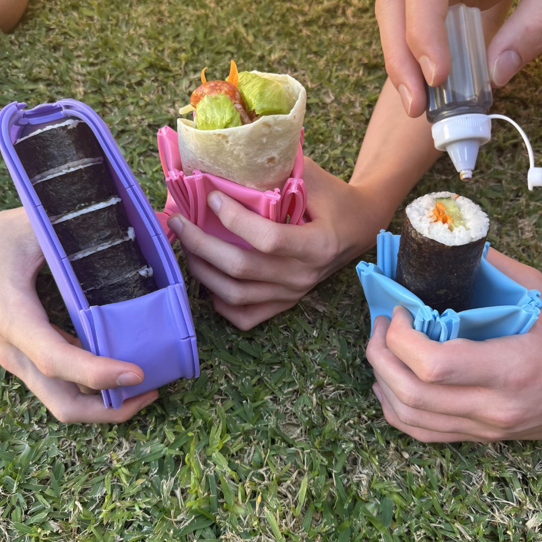 Hands holding colourful Wrap'd wrap holders containing sushi and fresh wraps on grass